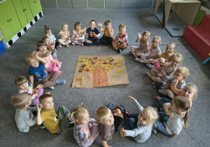Dzieci siedzą na dywanie wokół wspólnie wykonanej pracy plastycznej "Jesienne drzewo"