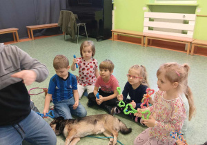 Dzieci trzymają w dłoniach zabawki dla psów. Na podłodze przed dziećmi leży pies