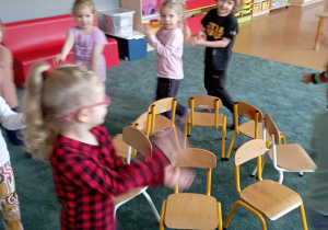 Dzieci podczas zabawy "Kto pierwszy?" chodzą między krzesłami, by jak ucichnie muzyka jak najszybciej usiąść na jednym z nich