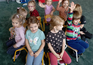 Dzieci siedzą na krzesłach ustawionych w kole na dywanie
