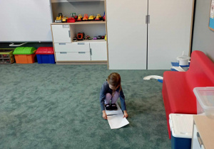 Dziewczynka kładzie kartkę A4 przed sobą na dywanie, by móc kontynuować dalszy spacer po kartkach