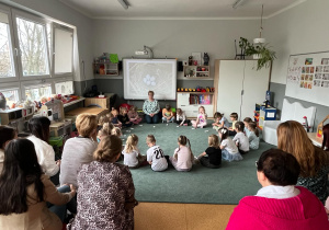 Dzieci i nauczycielka siedzą w kole na dywanie. Przed dziećmi leżą wykonane przez nich bociany