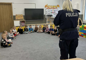 Dzieci w półkolu na dywanie siedzący i słuchający odwiedzającej Policjantki