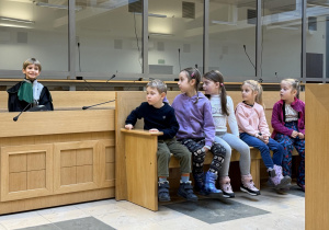 Dzieci obserwują i słuchają wypowiedzi chłopca wcielającego się w rolę obrońcy oskarżonego podczas symulowanej rozprawy sądowej