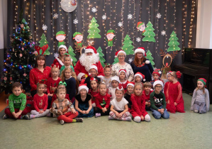 Dzieci z Grupy Sówki pozują do zdjęcia wraz z paniami, Mikołajem i Mikołajką na tle świątecznej dekoracji