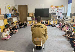 Dzieci w sali w kole na dywanie a przed nimi siedzący Strażak opowiadający o swoim zawodzie