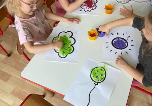 Dzieci wykonują prace plastyczną. Kropkują balony i chmury farbami za pomocą palców.