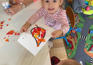 Dziewczynka przy stoliku pokazującą pracę plastyczną - liść wyklejony z bibuły