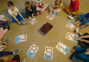 Sala przedszkolna. Dzieci siedzą w kole, na dywanie. Przed nimi karty pracy z wizerunkami cyfr od 1 do 9. Kształty cyfr utworzone są z sylwetek kasztanów. Na środku stoi prostokątne pudełko pełne kasztanów.