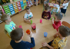 Sala przedszkolna. Dzieci siedzą przy stole, piją soki z kolorowych kubków. Na stole widać dzbanek z sokiem z buraka.