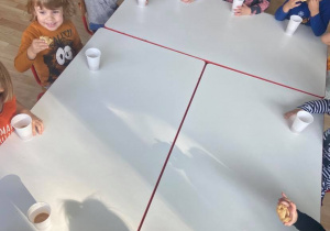 Dzieci przy stolikach pokazują ciasteczka oraz wyciśnięty sok z jabłka w kubkach