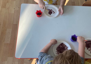 Dzieci przy stoliku malują papierowe talerzyki na brązowo