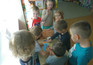 Sala przedszkolna. Dzieci zgromadzone przy stoliku pokazują książkę Misiowi Czytulkowi.