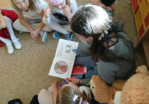 Sala przedszkolna. Dzieci siedzą na dywanie. Razem z nimi na dywanie siedzi kobieta czyta dzieciom książkę. Obok kobiety siedzi Miś Czytulek.