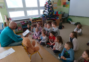 Sala przedszkolna. Dzieci siedzą na dywanie. Mężczyzna siedzi na krześle zwrócony do dzieci czyta im książkę. Na krześle obok siedzi Miś Czytulek.