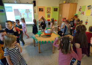 Sala przedszkolna. Dzieci tańczą dookoła stołu, na którym znajduje się zielona pacynka Zoom i plażowa piłka. W tle tablica interaktywna, na której widać obraz z piosenki ,,Ball"