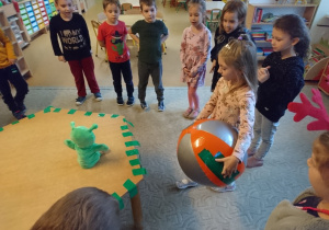 Sala przedszkolna. Dzieci ustawione w kole dookoła stołu, na którym znajduj się zielona pacynka Zoom. Dziewczynka przykleja do piłki zielone kawałki taśmy.