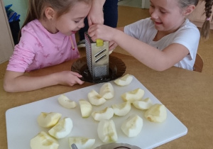 Sala przedszkolna. Dwie dziewczynki siedzą przy stole, trą na tarce jabłko, przed nimi na stole leżą pokrojone w ćwiartki jabłka..