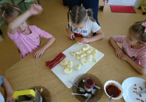 Sala przedszkolna. Trzy dziewczynki siedzą przy stole. Jedna z nich kroi nożem jabłko. Na stole leżą jabłka pokrojone w ćwiartki, kabanosy, ketchup i powidła owocowe.