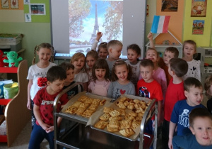 Sala przedszkolna. Dzieci ustawione dookoła wózka gastronomicznego, na którym znajdują się wykonane przez nich wypieki z ciasta francuskiego. Za dziećmi na tablicy interaktywnej obraz z wieżą Eiffla.