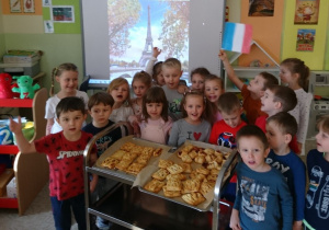 Sala przedszkolna. Dzieci ustawione dookoła wózka gastronomicznego, na którym znajdują się wykonane przez nich wypieki z ciasta francuskiego. Za dziećmi na tablicy interaktywnej obraz z wieżą Eiffla