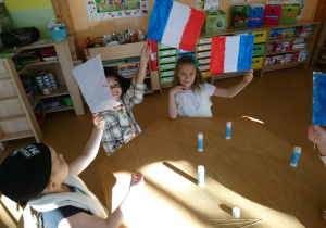 Sala przedszkolna. Dzieci siedzą przy stole machają wykonanymi przez siebie flagami Francji.