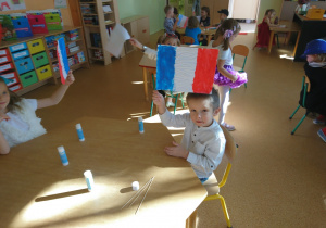 Sala przedszkolna. Dzieci siedzą przy stole machają wykonanymi przez siebie flagami Francji.