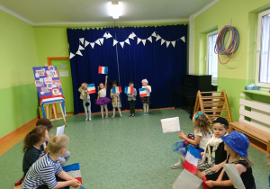 Sala gimnastyczna. Francuski pokaz mody. Grupa dzieci prezentuje stroje przygotowane wraz z rodzicami. Pozostałe dzieci siedzą na ławkach i obserwują pokaz.