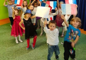 Sala gimnastyczna. Francuzki pokaz mody.Dzieci przebrane w stroje, ustawione w luźnej grupie machają flagami francuskimi.
