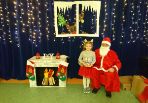 Sala gimnastyczna. Święty Mikołaj siedzi na fotelu, obok stoi dziewczynka w w białej bluzce i czerwonej spódnicy trzyma prezent. Z lewej strony udekorowany kominek. Na drugim planie, na granatowym tle dekoracja świąteczna, świecące lampki, okno wycięte z papieru w nim zaglądający renifer.