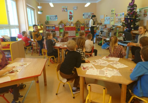 Sala przedszkolna. Dzieci siedzą przy stolikach i słuchają ciekawostek na temat czekolady, które opowiada im pani prowadząca zajęcia.