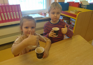 Sala przedszkolna. Chłopiec i dziewczynka siedzą przy stoliku, pokazują uniesione w górę kciuki po degustacji płynnej czekolady.