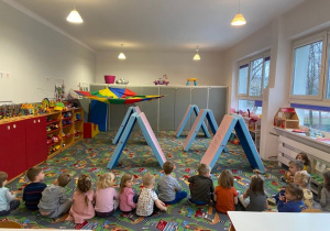 Dzieci w sali siedzą na dywanie. Przed nimi ustawiony tor przeszkód z materacy oraz namiot z chusty animacyjnej