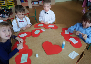 Sala przedszkolna. Dzieci siedzą przy stoliku i robią laurki w kształcie serca, dla Babci i Dziadka.