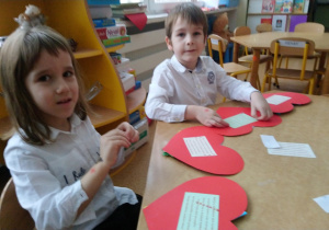 Sala przedszkolna. chłopiec i dziewczynka siedzą przy stoliku i robią laurki w kształcie serca, dla Babci i Dziadka.