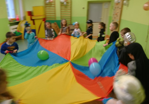 Sala gimnastyczna. Bal karnawałowy. Dzieci w przebraniach biorą udział w zabawie z chustą animacyjną, na chuście balony.