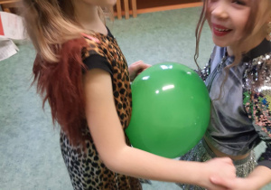 Sala gimnastyczna. Bal karnawałowy. Dwie dziewczynki w przebraniach tańczą w parze, trzymają balon między swoimi brzuchami.