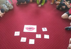Dzieci siedzą w kole na dywanie dookoła ilustracji przedstawiającej usta i wokół obrazków symbolizujących sytuacje, podczas których używamy życzliwych słów