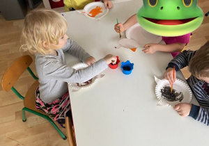 Dzieci przy stoliku przygotowują kota z talerzyków papierowych malowanych farbą