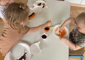 Dzieci przy stoliku przygotowują kota z talerzyków papierowych malowanych farbą