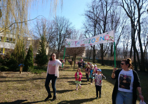 Ogród przedszkolny. Dzieci idą w pochodzie dookoła ogrodu i grają na instrumentach. Na czele pochodu dwie nauczycielki niosą transparent "Witaj Wiosno".