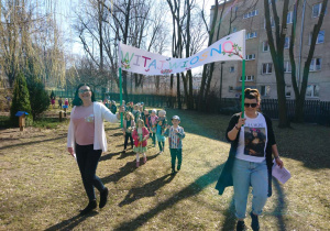 Ogród przedszkolny. Dzieci idą w pochodzie dookoła ogrodu i grają na instrumentach. Na czele pochodu dwie nauczycielki niosą transparent "Witaj Wiosno".