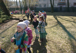 Ogród przedszkolny. Dzieci idą w pochodzie dookoła ogrodu i grają na instrumentach.