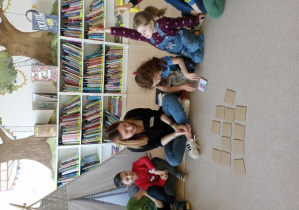 Dziewczynka siedząca obok Pani Bibliotekarski odkrywa jeden z obrazków w grze memo. Dziewczynka siedząca obok podnosi rękę do góry oznajmiając chęć bycia następną w kolejności do odkrywania kart