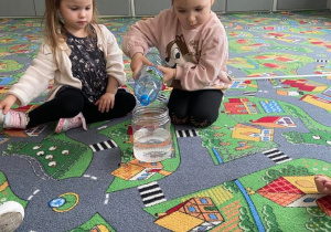Dwie dziewczynki siedzą na dywanie. Jedna z nich wlewa wodę do plastikowego pojemnika