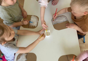 Dzieci przy stoliku malują kartoniki białą farbą