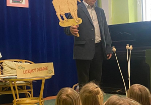 Autor książki recytuje dzieciom wiersze na sali gimnastycznej. W ręku drewnianą tabliczkę z mrówką. trzyma rekwizyt drewnianą tabliczkę z kozą