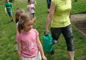 Ogród przedszkolny. Dzieci wraz z nauczycielką niosą wodę w konewce do podlewania roślin na grządce.