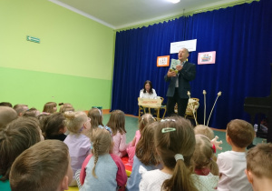 Sala gimnastyczna w przedszkolu. Dzieci siedą na widowni. Z uwagą patrzą na Eugeniusz Dolata prezentującego swoją książkę.