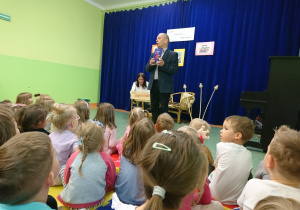 Sala gimnastyczna w przedszkolu. Dzieci siedą na widowni. Z uwagą patrzą na Eugeniusz Dolata prezentujacego swoją ksiązkę.